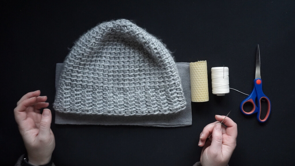 How to line a crochet beanie with fleece 1 Materials for lining a beanie, including the hat itself, the fabric beneath it, cotton thread, a beeswax candle, a needle and a pair of scissors.