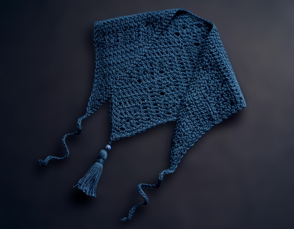 Dark blue triangle bandana folded with the ends over the front, with an intricate overall lace pattern, curly ties and a tassel with a bead attached to the point at the bottom, over a black background.