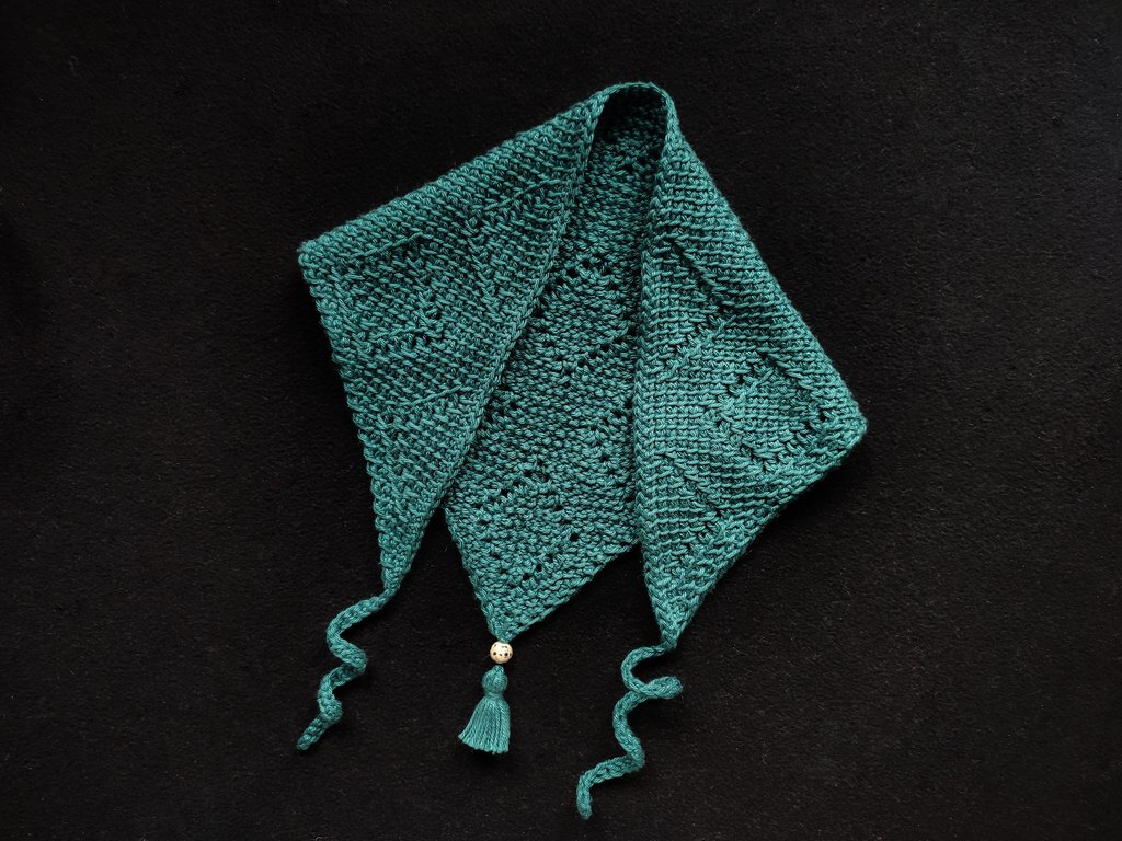 Green triangle bandana folded with the two ends towards the front, to create an almost square shape. The fabric has offset diamond shaped motifs made in lacy Tunisian crochet stitches and the two ties are curled towards the bottom. A small tassel with a bead is attached at the bottom.