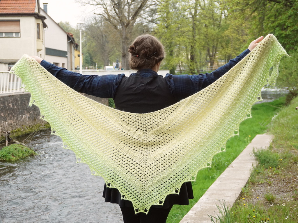 Light yellow to light green gradient Tunisian crochet shawl made in a rounded triangle shape, featuring surface stitches and edge stitches that create a zig zag along solid patterned slices, with other slices made up of lacy stitches. 