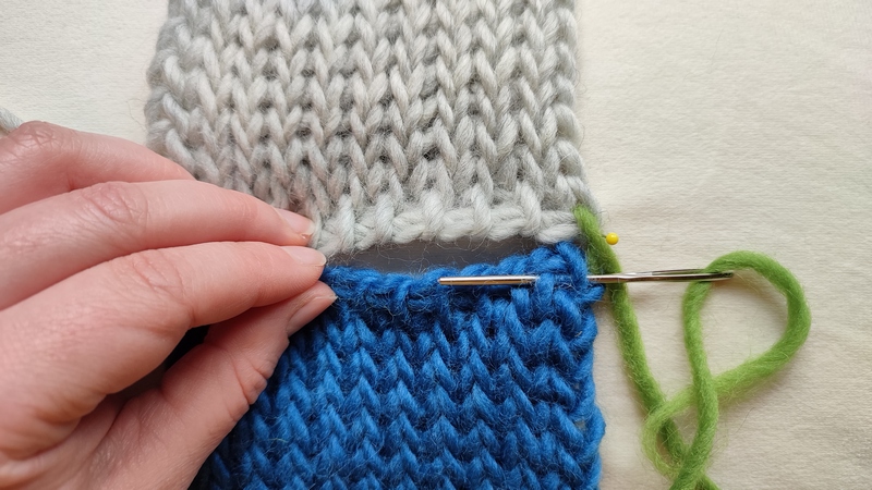 How to sew Tunisian knit stitch row to row 12 Close-up of two Tunisian crochet swatches being seamed together using a yarn needle and green yarn. One swatch is light gray and the other is blue. A hand holds the pieces in place while the needle is inserted behind the front vertical loop of the edge stitch and the back vertical bar of the first Tunisian knit stitch in the top row.