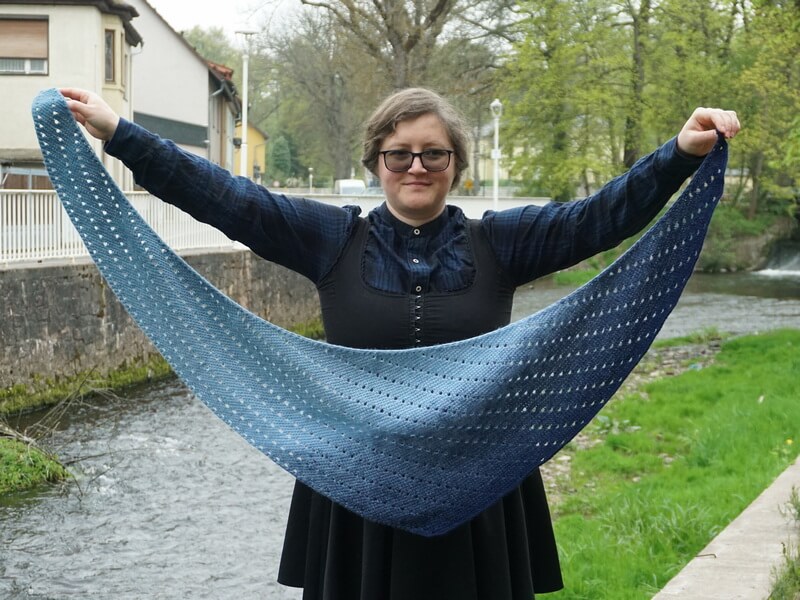 Tunisian crochet asymmetrical rounded triangle shawl tutorial 6 Light blue to dark blue asymmetrical rounded triangle shawl made in fingering weight yarn. The shape is more of a boomerang, with one edge longer than the other. There are rows of eyelets going across the fabric, in parallel to the longer side.