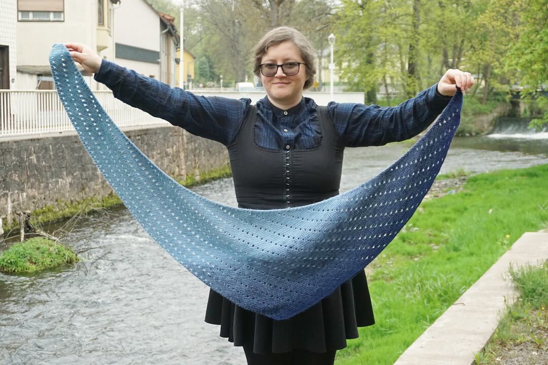 How to make different shawl shapes in Tunisian crochet 21 Light blue to dark blue asymmetrical rounded triangle shawl made in fingering weight yarn. The shape is more of a boomerang, with one edge longer than the other. There are rows of eyelets going across the fabric, in parallel to the longer side.
