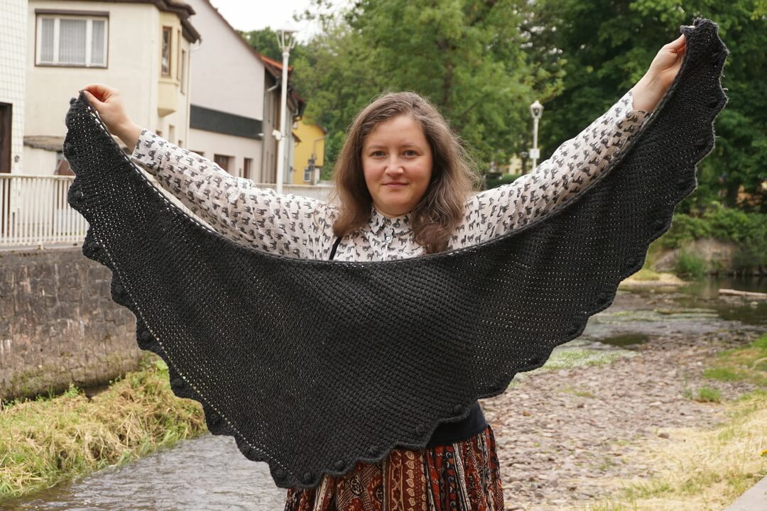 How to make different shawl shapes in Tunisian crochet 20 Dark gray asymmetrical rounded triangle shawl made in worsted weight yarn. The shape is more of a boomerang, with one edge longer than the other. There are scallops on the edge, with one popcorn stitch in the middle of each scallop.