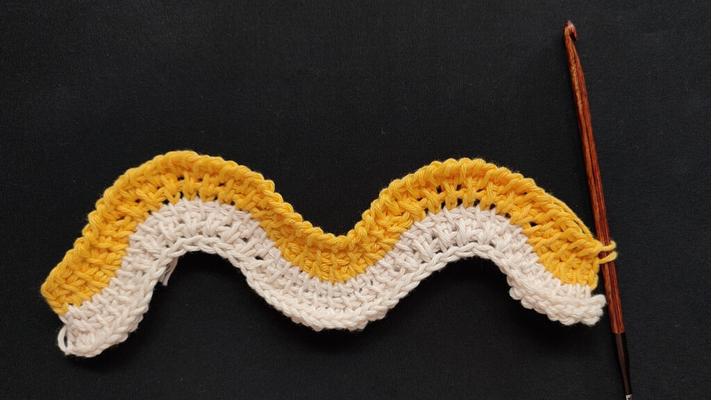 Feather and fan Tunisian crochet pattern for a washcloth 6 Six rows completed in the Tunisian crochet feather and fan washcloth, with the bottom three rows in a beige yarn and the next three rows in yellow. The wave is not as pronounced, as there are two plain rows between the rows with increases and decreases.