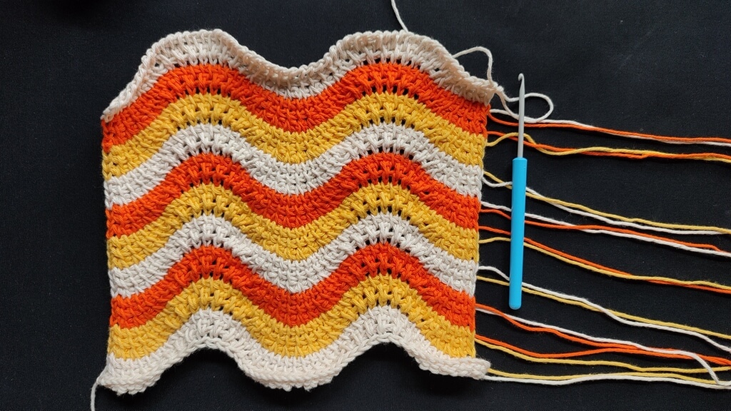 Feather and fan Tunisian crochet pattern for a washcloth 7 Finished fabric of the Tunisian crochet feather and fan pattern, with 10 stripes of 3 rows each, in alternating beige, yellow, orange colors. Unwoven ends are towards the right of the photo, under a small regular crochet hook.