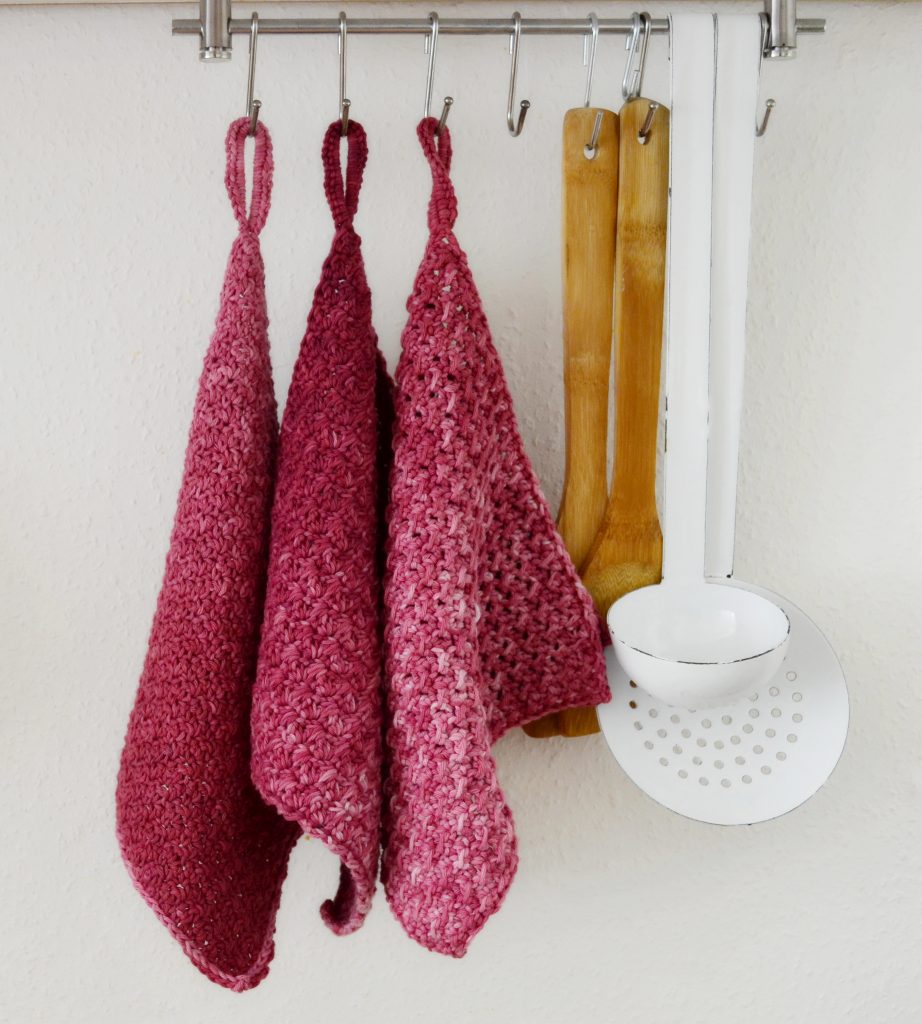 Three beautiful crochet washcloths on hooks, next to a strainer, a ladle and two bamboo spatulas